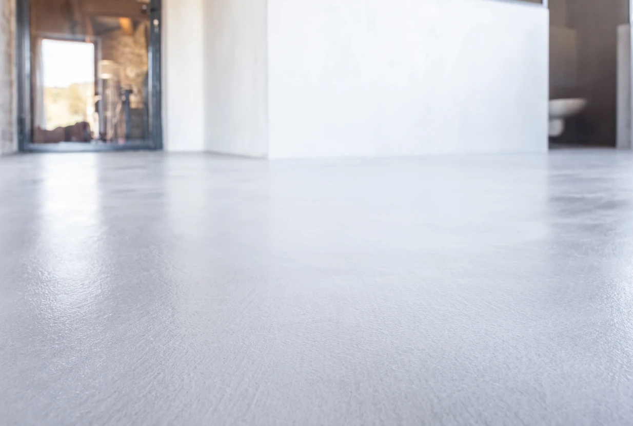 Professionnel en tenue de travail et équipements de sécurité (masque, gants) utilisant une ponceuse-polisseuse électrique sur une grande dalle de béton gris clair dans un intérieur, avec derrière lui plusieurs sections de sol montrant les différentes étapes de polissage, du béton brut au béton poli brillant et réfléchissant.