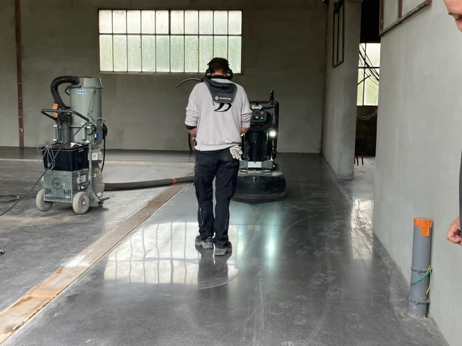 Plan rapproché d'une surface de béton poli luisant, montrant le résultat final du polissage avec une finition brillante et lisse, révélant les teintes naturelles du béton. Un artisan en tenue de travail professionnelle tenant une polisseuse ou une meuleuse angulaire au-dessus de la surface en cours de finition, avec des poussières de polissage visibles, démontrant le savoir-faire technique du travail de polissage. Environnement professionnel clair et bien éclairé mettant en avant la technicité et la qualité du résultat.