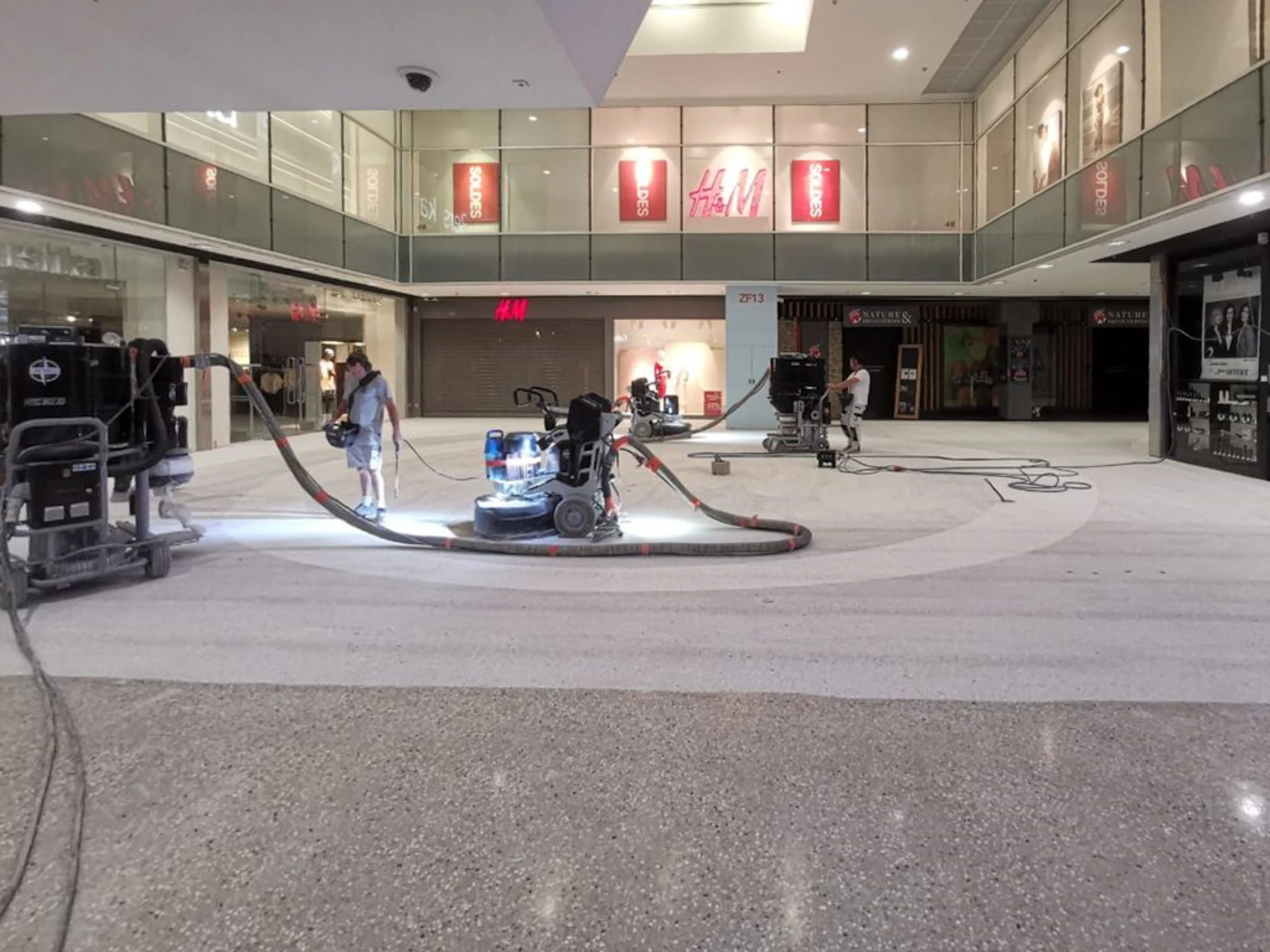 Opérateur en combinaison de travail grise, casque antibruit, gants, guidant une machine de polissage de sol béton (meuleuse planétaire) dans un local industriel vide